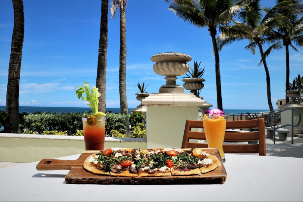 A scenic beachfront brunch setting featuring a beautifully crafted flatbread topped with fresh greens and tomatoes, served alongside two vibrant drinks—a Bloody Mary garnished with celery and a tropical cocktail adorned with a flower. The view includes palm trees, elegant stone balustrades, and the ocean in the background, creating a perfect backdrop for a relaxing and luxurious brunch experience.