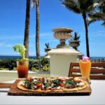 A scenic beachfront brunch setting featuring a beautifully crafted flatbread topped with fresh greens and tomatoes, served alongside two vibrant drinks—a Bloody Mary garnished with celery and a tropical cocktail adorned with a flower. The view includes palm trees, elegant stone balustrades, and the ocean in the background, creating a perfect backdrop for a relaxing and luxurious brunch experience.