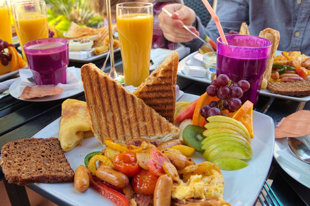 A vibrant brunch spread featuring a variety of colorful dishes, including toasted sandwiches, scrambled eggs, sausages, and an assortment of fresh fruits like grapes, oranges, kiwi, and apples. The table is adorned with glasses of fresh orange juice and a purple smoothie, creating a visually appealing and appetizing scene. The setting appears to be outdoors, with natural light highlighting the freshness of the food, making it an inviting meal in a relaxed environment.