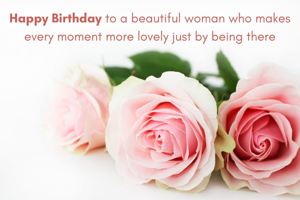 Elegant birthday greeting featuring three soft pink roses with lush green leaves resting on a white background. Above the flowers, in stylish brown-red font, the message reads: ‘Happy Birthday to a beautiful woman who makes every moment more lovely just by being there.’ The design conveys femininity, warmth, and grace, perfect for celebrating a special woman’s birthday