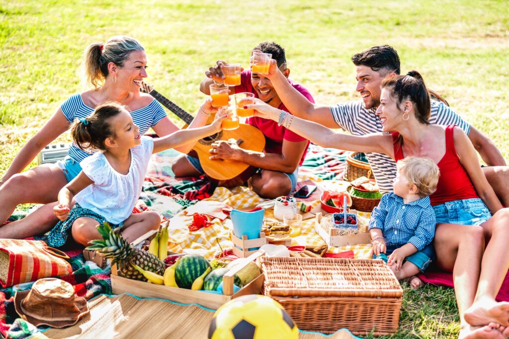 Joyful family picnic on a sunny day with adults and children toasting with juice, surrounded by vibrant blankets, fresh fruits, picnic baskets, and a guitar, creating a cheerful summer outdoor gathering atmosphere.