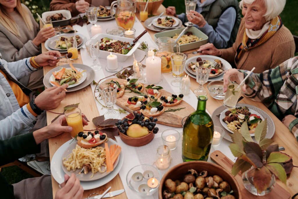 Family-style outdoor dinner gathering with assorted pasta dishes, bruschetta, roasted vegetables, fresh fruit, and candles on a rustic wooden table, surrounded by multi-generational guests enjoying a cozy autumn meal.