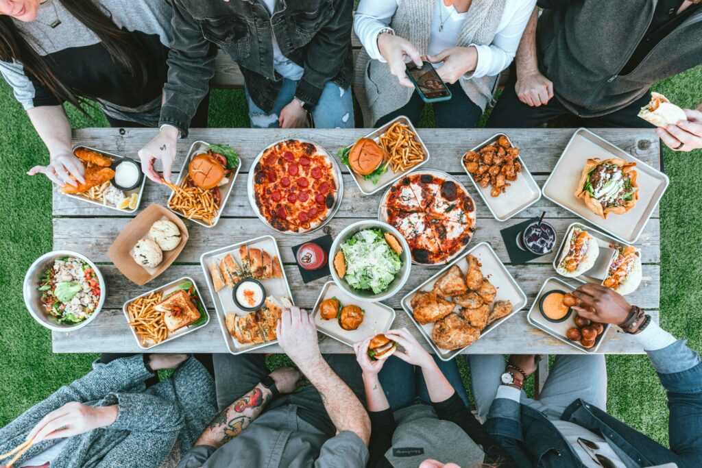 Overhead view of friends sharing a casual outdoor meal at a rustic wooden table filled with pizza, fries, burgers, salads, and dipping sauces, capturing a lively picnic or backyard gathering atmosphere.