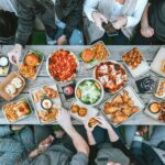 Overhead view of friends sharing a casual outdoor meal at a rustic wooden table filled with pizza, fries, burgers, salads, and dipping sauces, capturing a lively picnic or backyard gathering atmosphere.