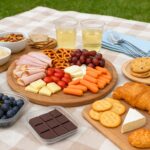 picnic setup for four people on a beige checkered blanket in a green park, featuring sliced baguette, pita bread, cheese cubes, deli meats, hummus cups, grapes, cherry tomatoes, cucumber sticks, baby carrots, pretzels, mixed nuts, brownies, and sparkling water bottles neatly arranged in trays and containers, with reusable cups, napkins, and a picnic basket in the background