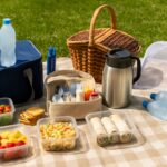 Realistic picnic setup on a beige checkered blanket with reusable containers of pasta salad, fresh strawberries, mango slices, green salad with cherry tomatoes, wraps, crackers, frozen water bottles, navy cooler bag, wicker picnic basket, stainless steel thermos, utensil and condiment caddy, bug spray, and mesh food cover arranged on lush green grass for an organized, time-saving outdoor meal.