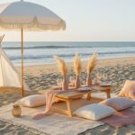 Elegant beach picnic scene with a white fringed umbrella, small wooden table, blush pink pampas grass arrangements, neatly set plates and glasses, cozy cream and blush cushions, and a white teepee on soft sand by the shoreline.