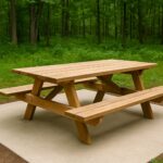 A wooden ADA-compliant picnic table on a concrete pad in a forest park setting. The table features extended space at one end without a bench, providing wheelchair accessibility. Surrounded by lush green grass and tall trees, the table is designed with clear ground access for inclusive outdoor use.