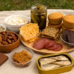 Keto picnic snacks displayed on a blanket outdoors with cheese crisps, salami, nuts, olives, beef jerky, and canned sardines as low-carb, no-refrigeration options.