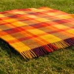 Bright plaid wool picnic blanket with bold yellow, orange, and red stripes accented by subtle purple and brown tones, lying on a sunny grassy field with visible natural texture and soft daylight shadows.