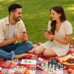 Young couple enjoying a picnic date on a red and white checkered blanket in a sunny park, playing UNO cards and surrounded by picnic essentials—cheese, bread, fruit, wine glasses, a wicker basket, sketchbook, and chess set—capturing the fun and relaxed mood of picnic date games.