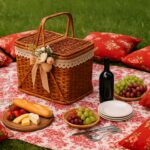 Vintage wicker picnic basket decorated with lace and flowers, placed on a patterned picnic blanket outdoors. Surrounded by eight red floral cushions, the setup includes picnic essentials: a wooden board with bread, cheese, and strawberries, bowls of grapes, a wine bottle, glassware, stacked plates, and cutlery. The arrangement reflects vintage picnic basket decorating ideas with a rustic and elegant style.